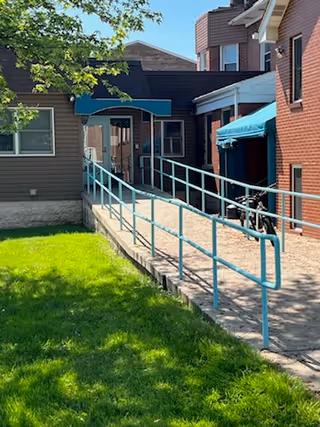 Outdoor view of a building entrance with a paved walkway and a blue handrail ramp leading to a covered doorway. There is green grass on the left side and parts of the building are made of brick and siding. A bicycle is parked near the entrance under a blue awning.