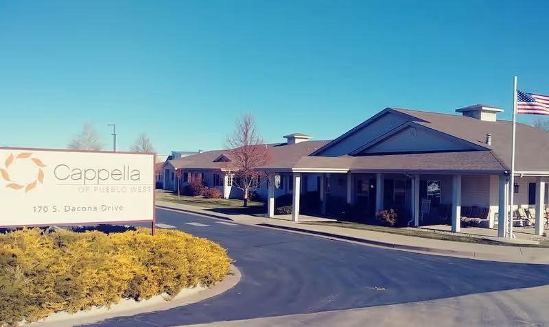 Exterior view of Cappella at Pueblo West senior living facility with a clear blue sky, a sign displaying the facility name and address, a driveway, landscaped bushes, and an American flag on a flagpole.