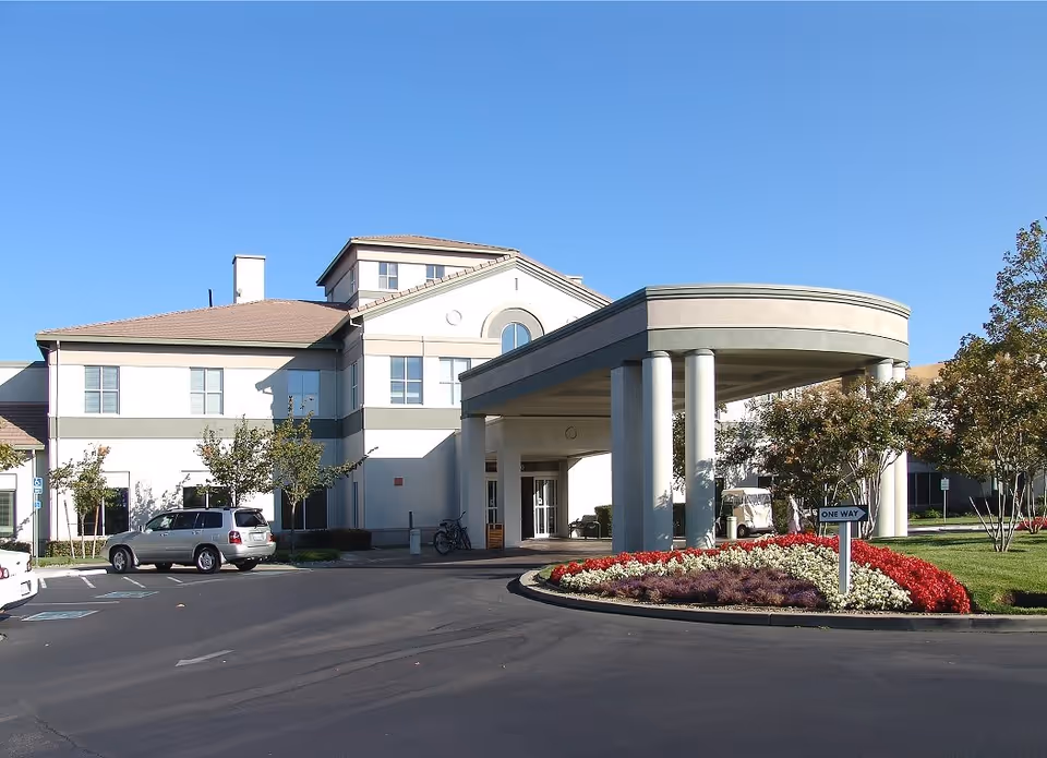 Exterior view of a senior living facility named Paradise Valley Estates with a covered entrance supported by columns, a landscaped roundabout with red and white flowers, several trees, and parked cars under a clear blue sky.