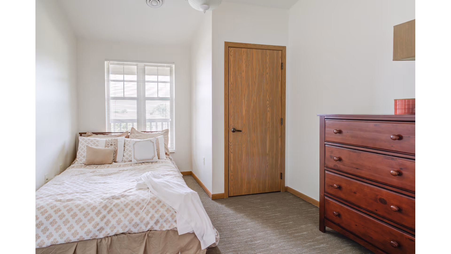 Small bedroom with a bed by a window, a wooden door, and a dark wood dresser.