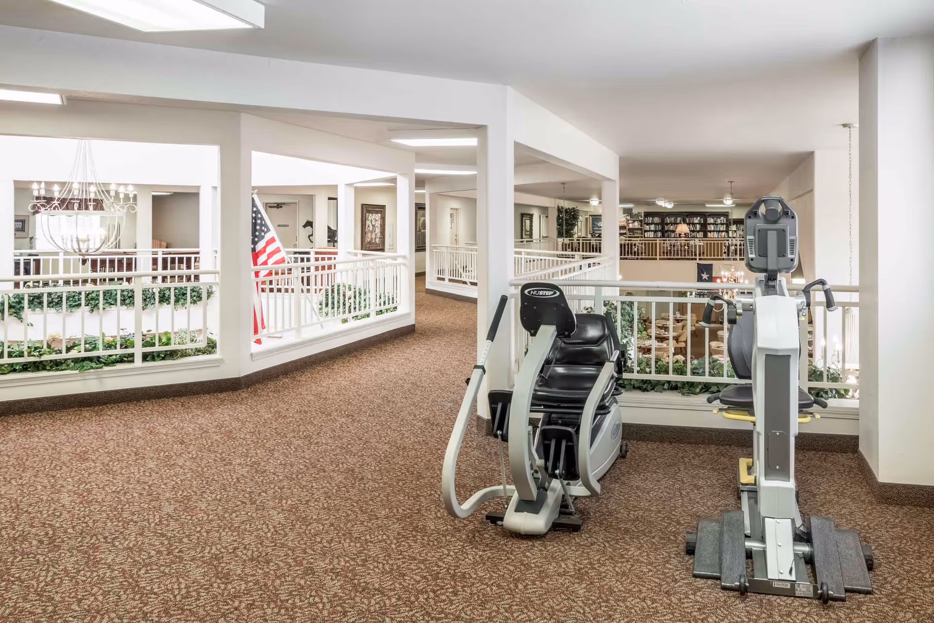 Interior view of a senior living facility hallway with exercise equipment including a NuStep machine. The hallway has carpeted floors, white railings, and overlooks a lower level with seating and bookshelves. An American flag is visible near the railing.