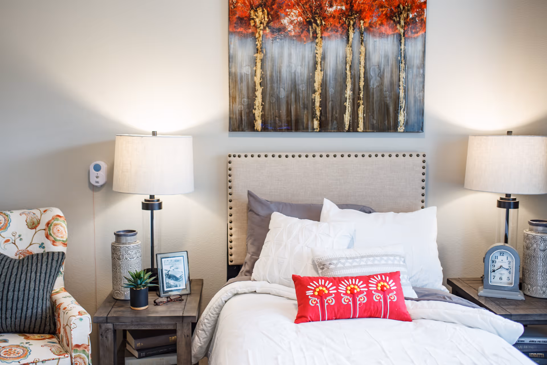 A cozy bedroom featuring a bed with a beige upholstered headboard adorned with nailhead trim. The bed is made with white and gray pillows, including a decorative red pillow with floral embroidery. On either side of the bed are wooden nightstands with matching table lamps, decorative vases, a small plant, a framed photo, and a clock. To the left, there is a floral-patterned armchair with a dark gray cushion. Above the bed hangs a large painting of trees with red foliage.