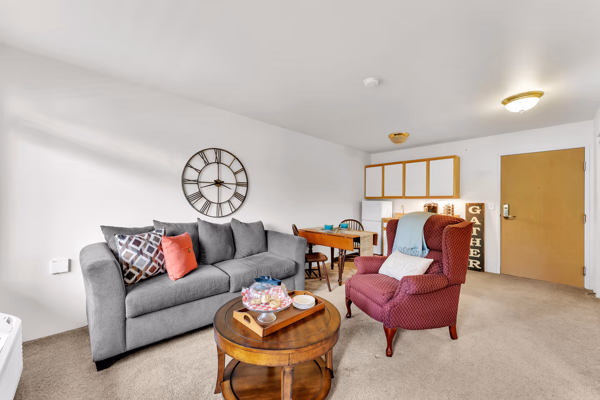 A cozy living room area featuring a gray sofa with patterned and solid pillows, a round wooden coffee table with a tray holding plates and a covered dish, and a red upholstered armchair with a light blue throw and a decorative pillow. Behind the seating area is a small dining table with chairs, a compact kitchenette with white cabinets, a refrigerator, and a coffee maker. A large decorative wall clock hangs on the white wall, and a wooden door is visible in the background.