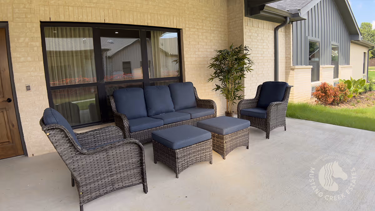 Outdoor patio area with wicker furniture including a sofa, two armchairs, and two ottomans with dark blue cushions, next to a beige brick wall and a window. There is a potted plant in the corner and some landscaping with grass and bushes nearby.