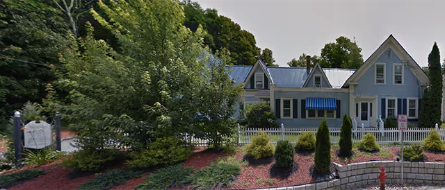 Exterior view of a light blue house with a white picket fence, surrounded by trees and landscaped bushes. The house has a metal roof, multiple dormer windows, and a blue striped awning over one window. There is a stone retaining wall and a fire hydrant in front of the property.