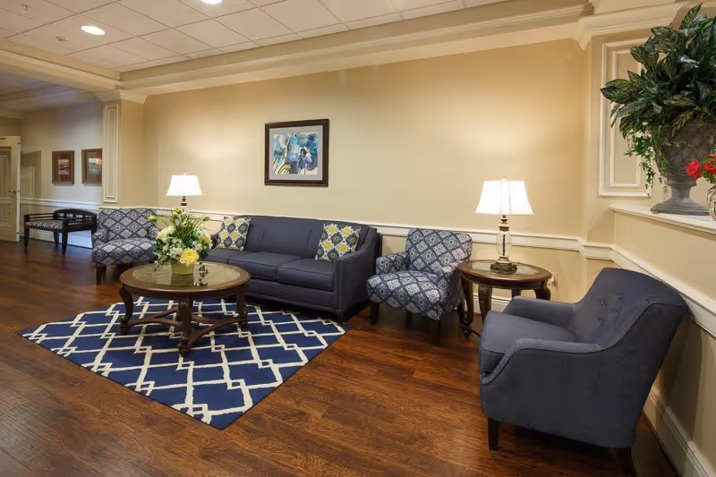 A cozy living room area with a dark blue sofa, two patterned armchairs, and a solid blue armchair arranged around a wooden coffee table with a floral centerpiece. There are two side tables with lamps, a blue and white patterned rug on a wooden floor, and a framed floral painting on the beige wall.