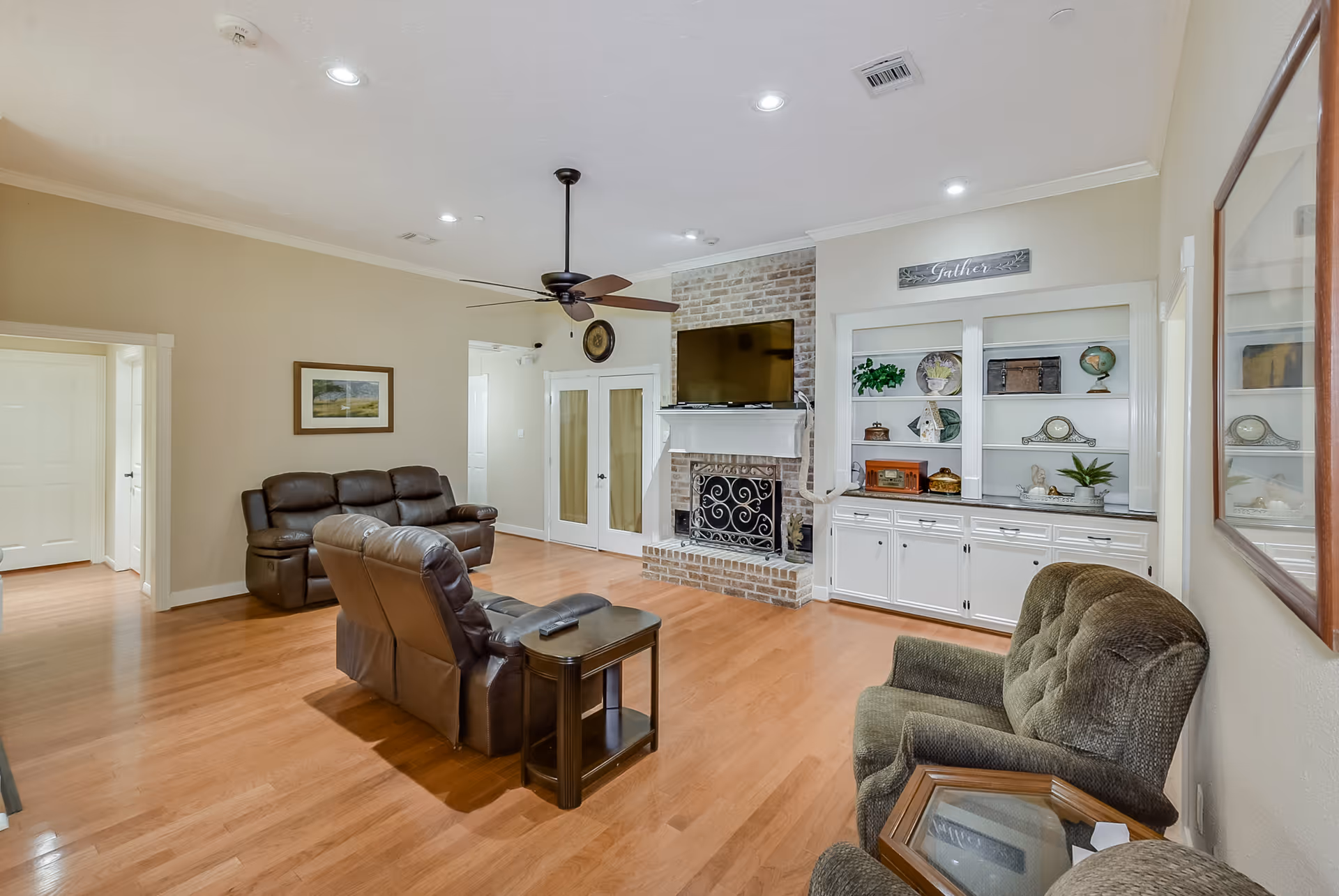 A spacious living room with wooden flooring, beige walls, and recessed ceiling lights. The room features a ceiling fan, a brick fireplace with a mounted flat-screen TV above it, and built-in white shelves decorated with various items. There are two brown leather recliners, a green upholstered chair, and a small wooden side table. A framed picture hangs on the wall, and double doors with glass panels are visible in the background.