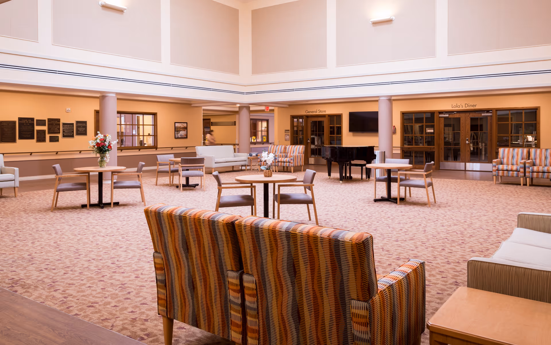 Spacious and well-lit common area with patterned armchairs, round tables with chairs, a grand piano, and a few sofas. The walls are painted beige with large windows and doors labeled 'General Store' and 'Lola's Diner'. There are flowers on some tables and plaques on the wall.