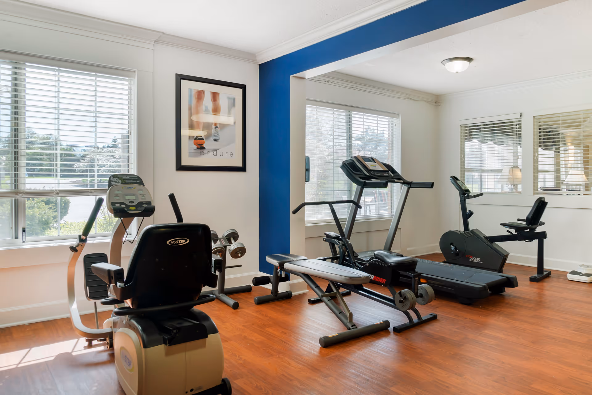 A bright fitness room with wooden flooring featuring exercise equipment including a recumbent bike, a treadmill, a rowing machine, and a stationary bike. Large windows with white blinds allow natural light to fill the space. A framed motivational poster with the word 'endure' hangs on the wall.