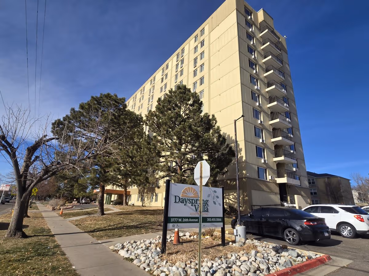 Tall multi-story Dayspring Villa senior living building with a front sign, trees, sidewalk, and parked cars under a blue sky.
