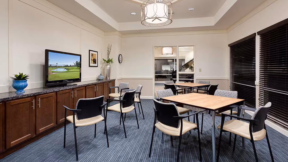 A well-lit common room with several tables and chairs arranged for group seating. There is a flat-screen TV mounted on a long wooden cabinet against the wall, with decorative plants and framed artwork nearby. The room has large windows with dark blinds and a modern ceiling light fixture.