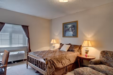 A cozy bedroom in a senior living facility featuring a wooden bed with a brown comforter and pillows, two bedside tables each with a lamp, a framed painting above the bed, a window with blinds and curtains, an air conditioning unit below the window, and a patterned armchair with an ottoman.