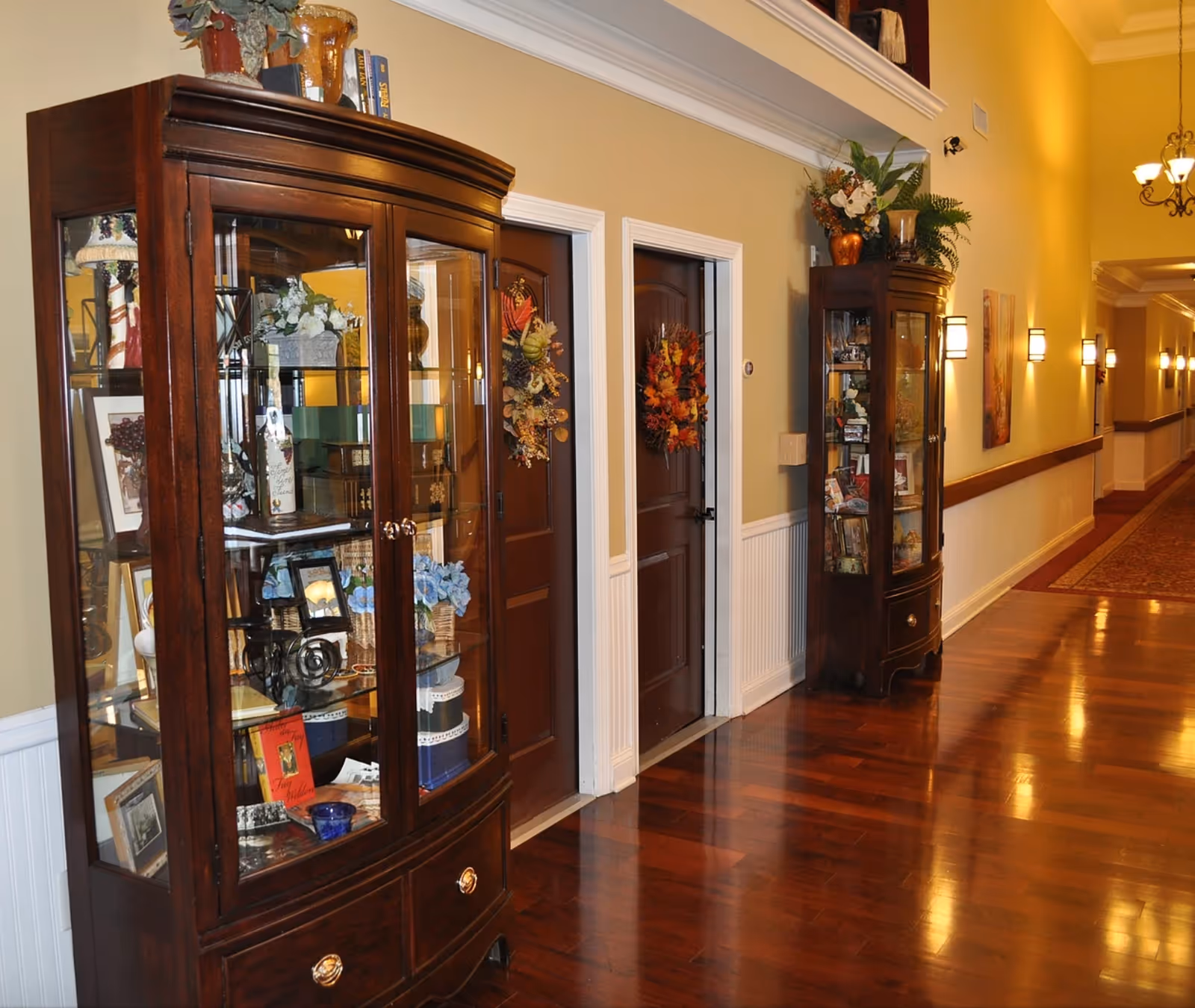 A well-lit hallway with polished wooden floors and beige walls featuring two dark wooden display cabinets filled with decorative items and books. Two closed wooden doors with autumn-themed wreaths hang on them. Wall sconces provide warm lighting along the corridor.