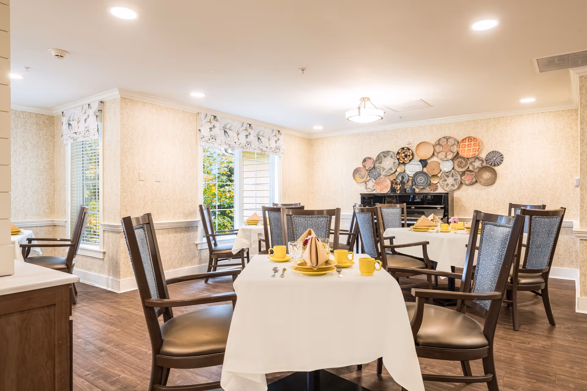 A bright dining room with several tables covered in white tablecloths, each set with yellow cups, plates, and folded napkins. The room has wooden chairs with cushioned seats and backs, floral-patterned window shades, and a decorative wall display of woven baskets above a black piano. The floor is wooden, and natural light comes through the windows.