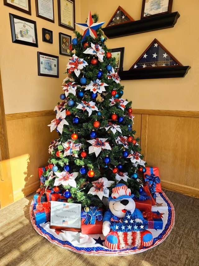 A decorated Christmas tree with red, white, and blue ornaments and large white flowers with red and blue accents. The tree is topped with a star in red, white, and blue colors. Around the base of the tree are wrapped presents in red and blue wrapping paper and a patriotic-themed teddy bear holding an American flag. The tree skirt is decorated with stars and stripes in red, white, and blue. The tree is placed in a corner with framed certificates and folded American flags displayed on shelves on the wall behind it.