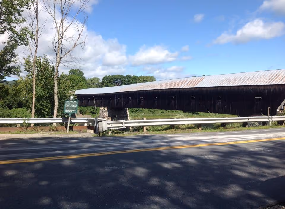 A long covered wooden bridge runs alongside a two-lane road with guardrails under a blue sky.