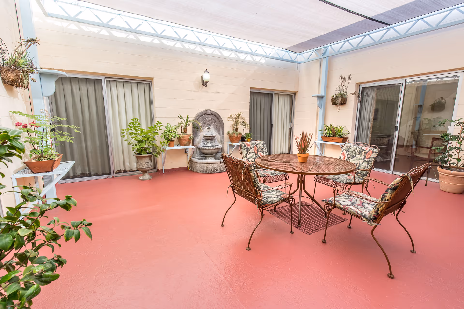 Indoor courtyard with a red floor, a round glass-top table with four cushioned metal chairs featuring floral patterns, several potted plants on shelves and the floor, a wall-mounted water fountain, and sliding glass doors leading to adjacent rooms. The area is covered with a translucent roof allowing natural light.