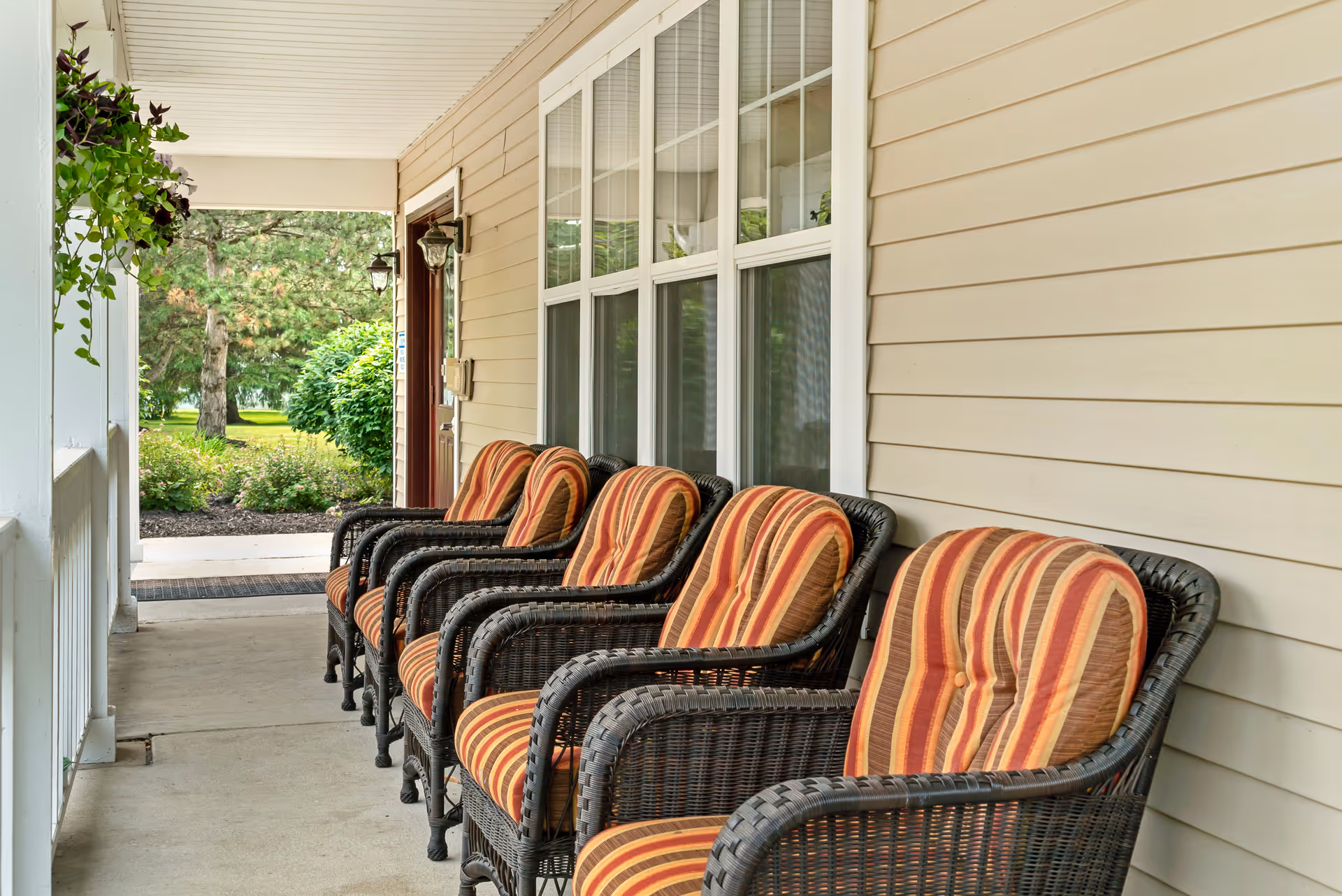 A covered porch with a row of five wicker chairs featuring striped orange and brown cushions. The porch has beige siding, white-framed windows, a hanging plant, and outdoor lantern-style lights. Green bushes and trees are visible in the background.