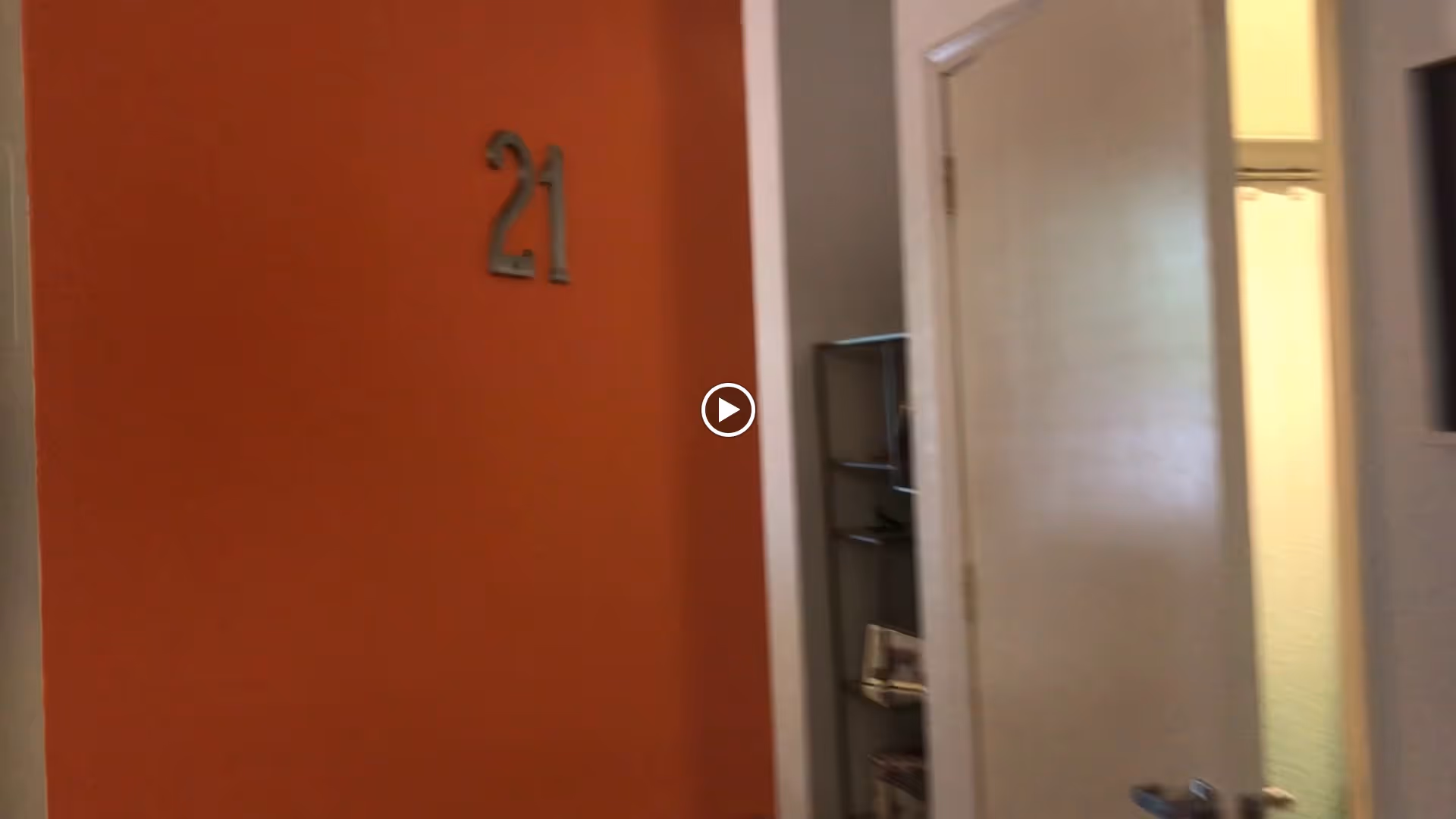 Interior view of a hallway with an orange wall displaying the number 21, a white door slightly ajar, and a metal shelving unit with some items on it.