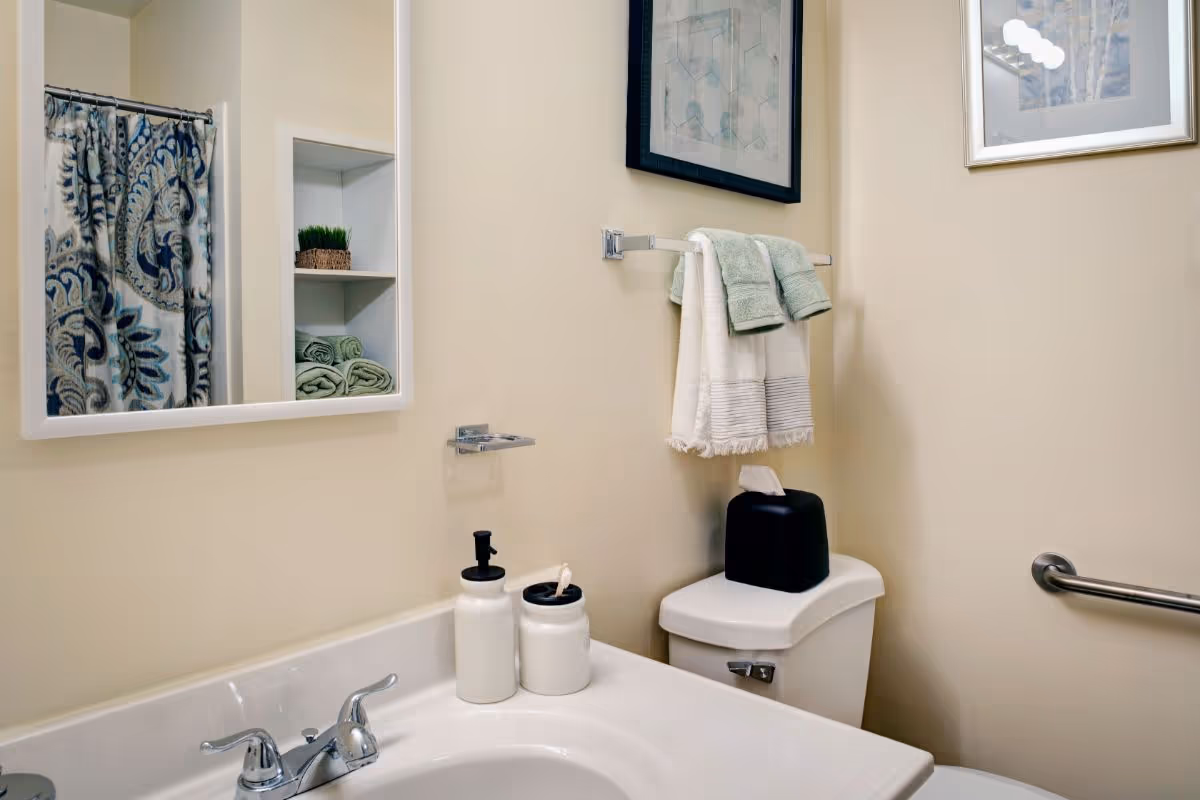 A clean bathroom with a sink and mirror, towels on a rack above the toilet, and a patterned shower curtain visible.