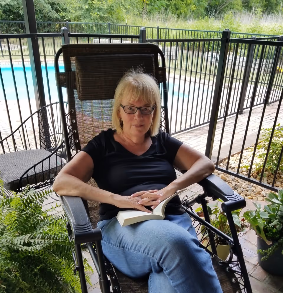 A woman with blonde hair and glasses sits comfortably in a reclining outdoor chair on a patio. She is wearing a black shirt and blue jeans, holding an open book in her lap. Behind her is a black metal fence enclosing a swimming pool, with greenery and plants around the patio area.