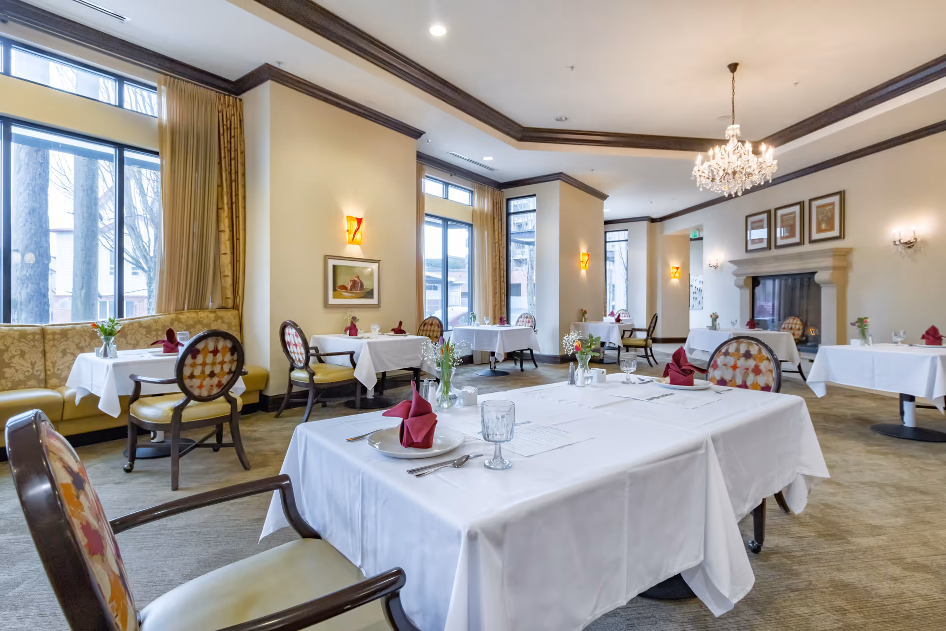 A bright and elegant dining room with multiple tables covered in white tablecloths, each set with plates, glasses, silverware, and folded red napkins. The room features large windows with beige curtains, upholstered chairs with patterned backs, a fireplace with framed artwork above it, and soft wall sconces providing warm lighting.