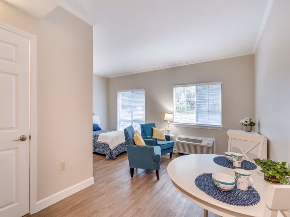 A bright and cozy senior living apartment featuring a small round dining table set with two blue placemats, cups, and bowls. Two blue armchairs with yellow pillows are positioned near a window with blinds, next to a white lamp and an air conditioning unit. A bed with blue and white bedding is partially visible in the background. The room has light-colored walls and wood flooring.