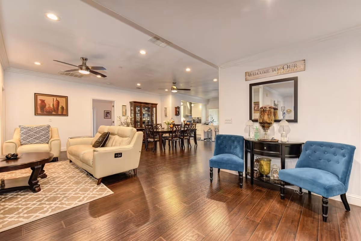 A spacious and well-lit living room area with wooden flooring, featuring a cream-colored sofa and armchair around a wooden coffee table on a patterned rug. Two blue upholstered chairs flank a black console table with decorative items and a mirror above it. In the background, there is a dining area with a wooden dining table and chairs, and a kitchen area further back. The walls are white with framed artwork and a sign that reads 'Welcome to Our Home'. Ceiling fans and recessed lighting are visible on the ceiling.