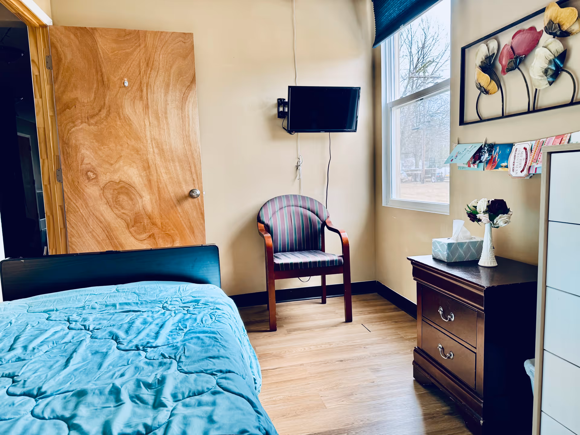 A small bedroom with a wooden door, a bed with a blue comforter, a striped armchair, a wall-mounted TV, a window with a view outside, a wooden nightstand with a tissue box and a vase of flowers, and a decorative wall art featuring colorful flowers.