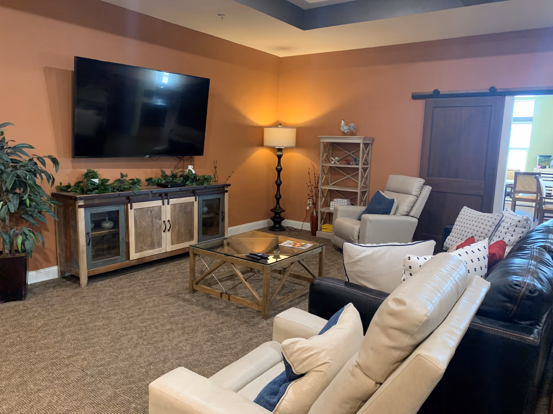 A cozy living room with a large flat-screen TV mounted on an orange wall above a wooden cabinet decorated with green plants. The room features a glass coffee table, several cushioned armchairs and a leather sofa with multiple pillows. A tall floor lamp and a wooden shelf with decorative items are in the corner. A sliding wooden door leads to another room with a dining table and chairs visible.