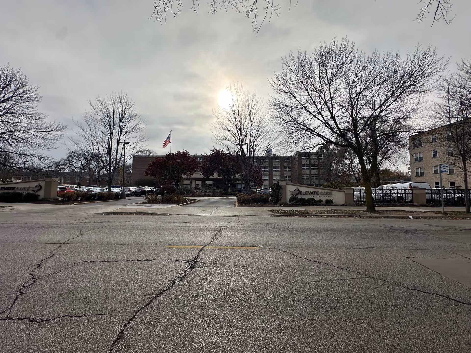 Exterior view of Elevate Care Chicago North facility with a parking lot in front, leafless trees, and an American flag flying near the entrance on a cloudy day.