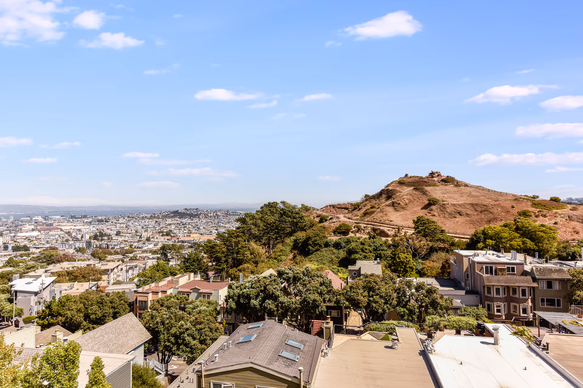 View of residential rooftops and a large grassy hill with the cityscape and blue sky in the background.