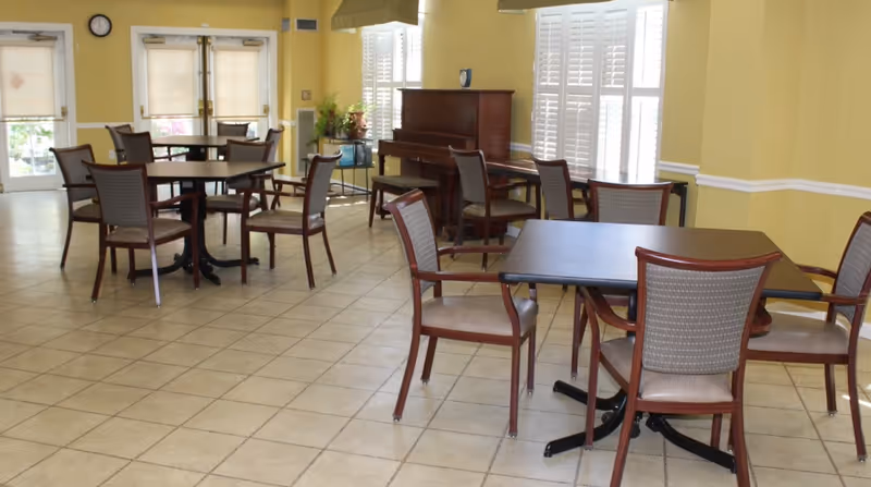 A bright dining room with several square tables and wooden chairs arranged neatly on a tiled floor. Large windows with white shutters allow natural light to fill the room. A piano is positioned against one wall, and there are plants and decorative items near the windows.