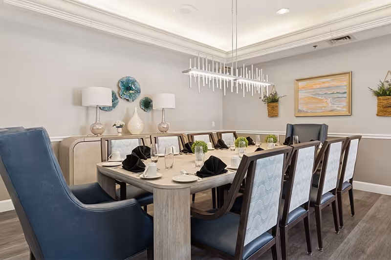 A well-lit dining room with a rectangular wooden table set for eight people. The table is arranged with black folded napkins, white plates, cups, and glasses. There are two large blue armchairs at the ends of the table and six wooden chairs with patterned upholstery along the sides. The room features a modern chandelier, decorative wall art including a beach painting and three blue-green ceramic wall pieces, two table lamps on a sideboard, and two hanging plants on the walls.