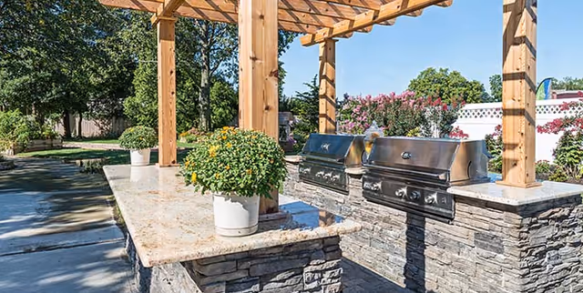 Outdoor grilling area with two stainless steel grills set into a stone countertop under a wooden pergola, surrounded by greenery and flowering plants.