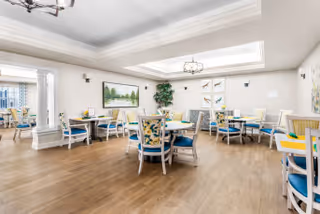 A bright dining room with multiple tables and chairs arranged neatly. The chairs have blue cushions and floral-patterned backs. The room features wooden flooring, light-colored walls, decorative wall art, and a large skylight allowing natural light to fill the space. There is a chandelier hanging from the ceiling and a large plant in one corner.