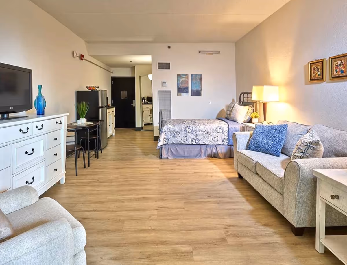 Studio-style assisted living room with a bed, sofa, TV on a dresser, small table, and wood flooring.