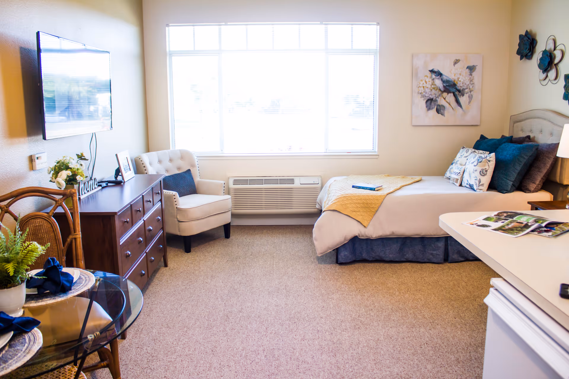 Bright furnished studio room with a bed, dresser, armchair, TV and small dining table beneath a large window.