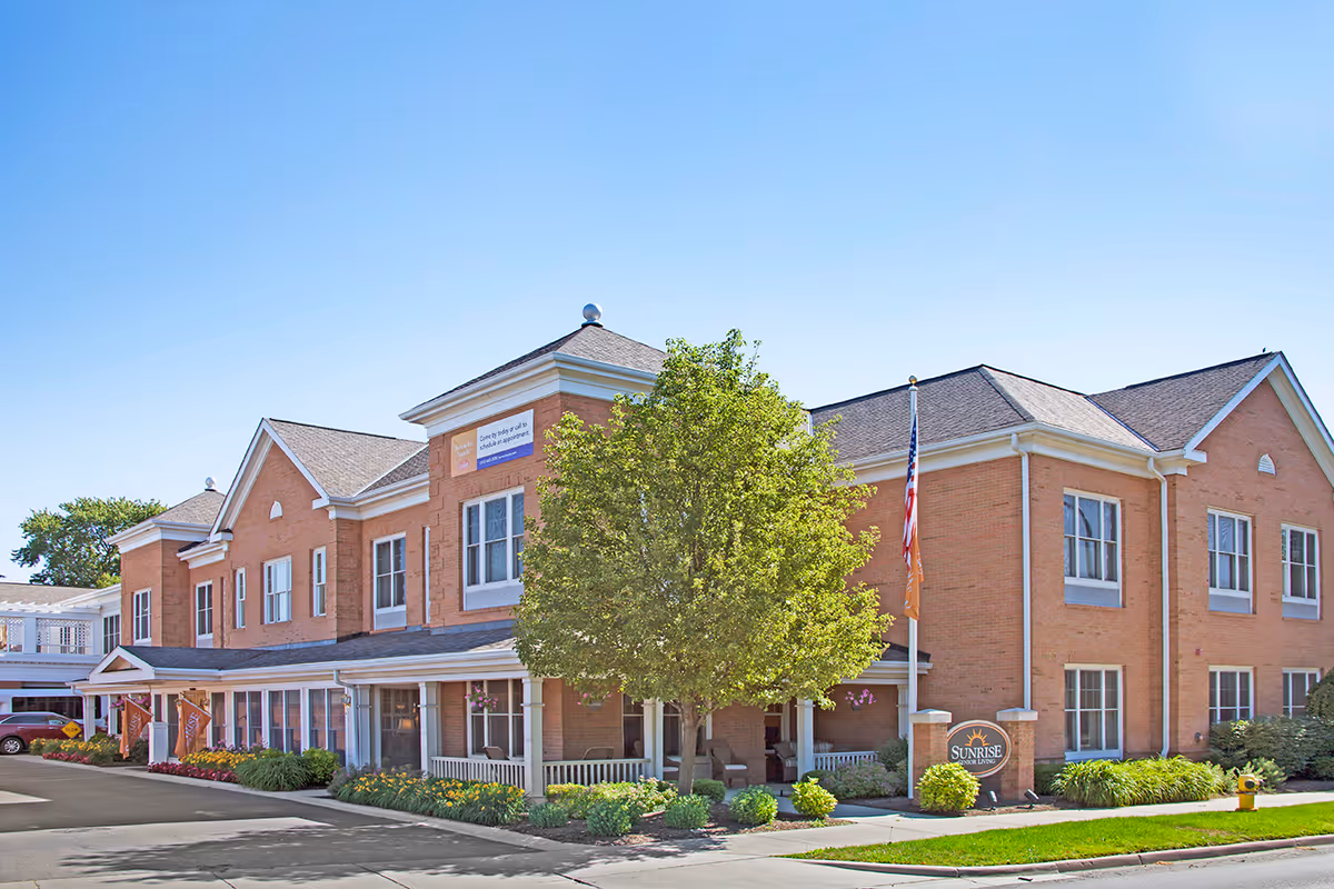 Exterior view of a two-story brick building with a porch, a tree in front, and a sign that reads Sunrise Senior Living. The building has multiple windows and a flagpole with an American flag and an orange flag. The sky is clear and blue.