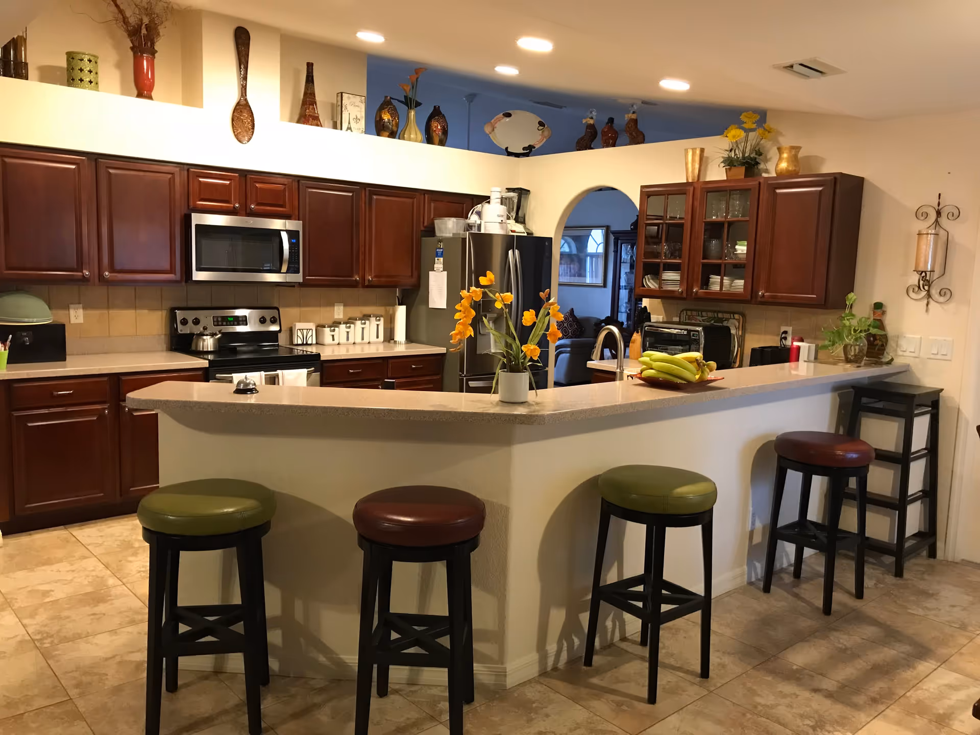 A spacious kitchen with dark wooden cabinets, a stainless steel microwave and stove, and a refrigerator. There is a long beige countertop with four bar stools, two with green seats and two with brown seats. The countertop has a vase with yellow flowers and a plate with bananas. Decorative items are displayed on a high shelf above the cabinets. The kitchen opens into a living area visible through an arched doorway.