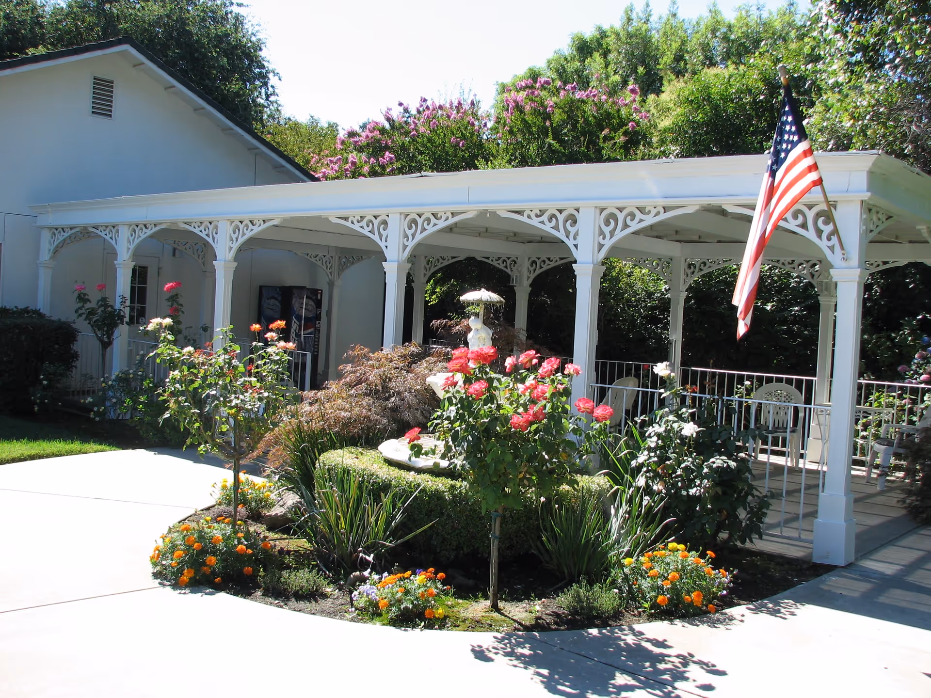 White decorative covered porch with an American flag and colorful flower beds in front of a single-story building.