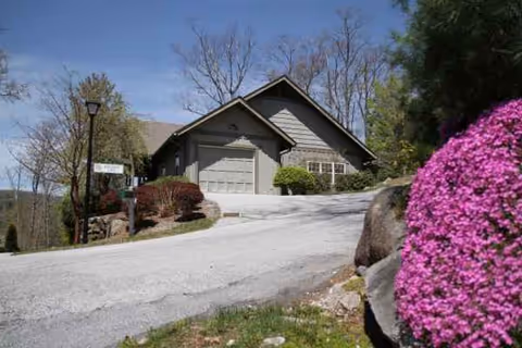 A gray, gabled senior living building sits at a curved driveway with pink azaleas.