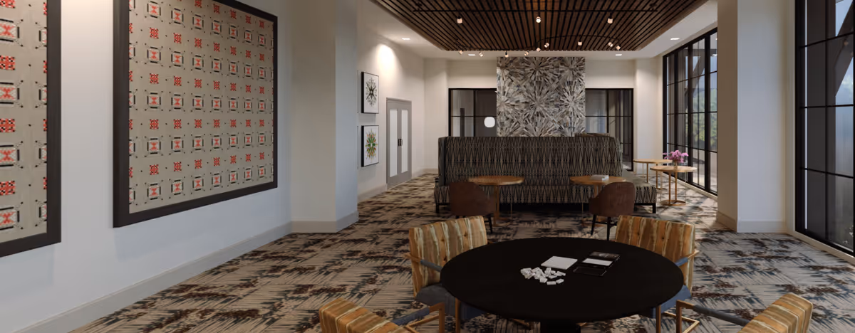 Communal lounge with round tables and upholstered seating, patterned carpet, artwork on the walls, and large floor-to-ceiling windows.