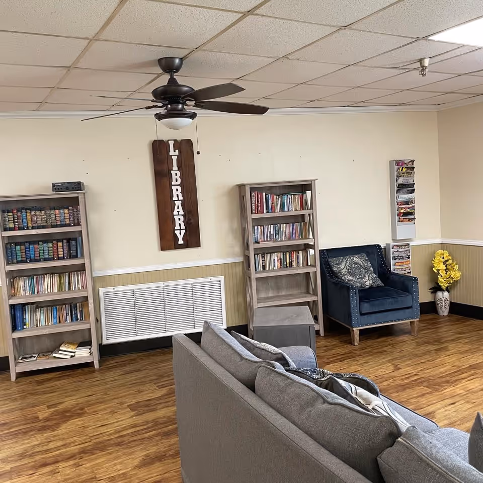 A cozy library corner in a senior living facility with two wooden bookshelves filled with books, a dark blue armchair with a patterned cushion, a gray sofa, a wooden floor, and a ceiling fan. A wooden sign with the word 'LIBRARY' hangs on the wall between the bookshelves. There is also a magazine rack on the wall and a vase with yellow flowers in the corner.