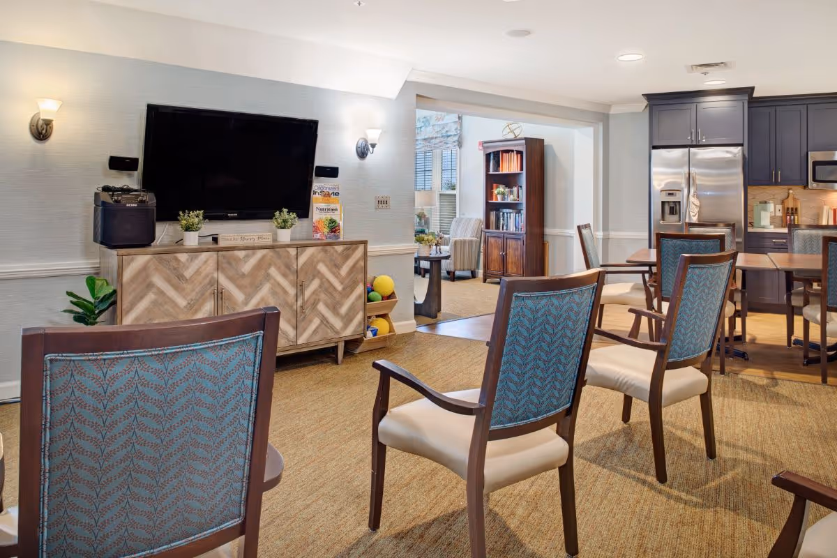 A cozy common area in a senior living facility featuring several wooden chairs with blue patterned backs arranged facing a wall-mounted flat screen TV above a wooden cabinet. The room has beige carpeting and soft lighting from wall sconces. In the background, there is a kitchen area with dark cabinets, a stainless steel refrigerator, and a microwave. Beyond the kitchen, another seating area with armchairs and a bookshelf is visible.
