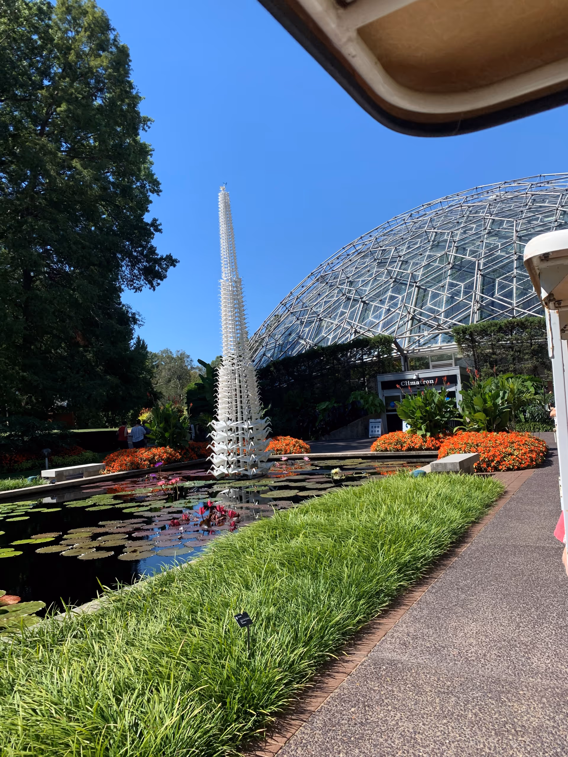 Outdoor garden area with a pond filled with lily pads and a tall white sculpture rising from the water. Surrounding the pond are green grass, orange flowers, and various plants. In the background, there is a large glass dome structure under a clear blue sky.