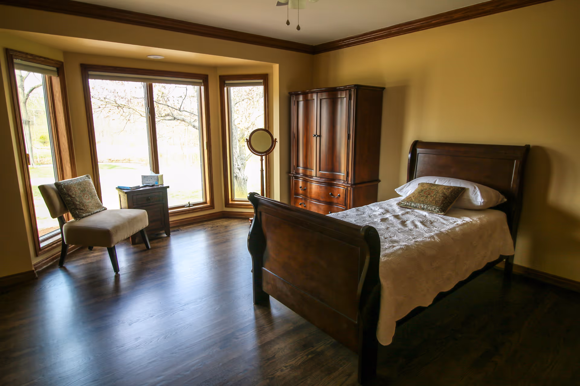 A cozy bedroom with a wooden bed frame, white bedding, and a decorative pillow. The room features dark hardwood floors, a wooden armoire, a small wooden nightstand, a cushioned chair with a pillow, and large bay windows letting in natural light with a view of trees outside.