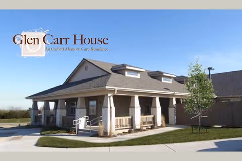 Exterior view of Glen Carr House, a single-story building with a covered porch, columns, and a sloped roof with dormer windows. There is a small tree and a paved walkway in front of the building under a clear blue sky.
