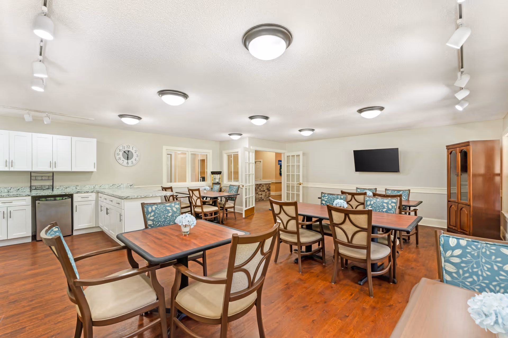 A bright and clean dining room with several wooden tables and chairs, some with floral patterned cushions. The room features a kitchenette with white cabinets, a granite countertop, and a small dishwasher. There is a wall clock above the kitchenette, a flat-screen TV mounted on the wall, and a wooden cabinet in the corner. The floor is hardwood, and the ceiling has multiple round light fixtures and track lighting.