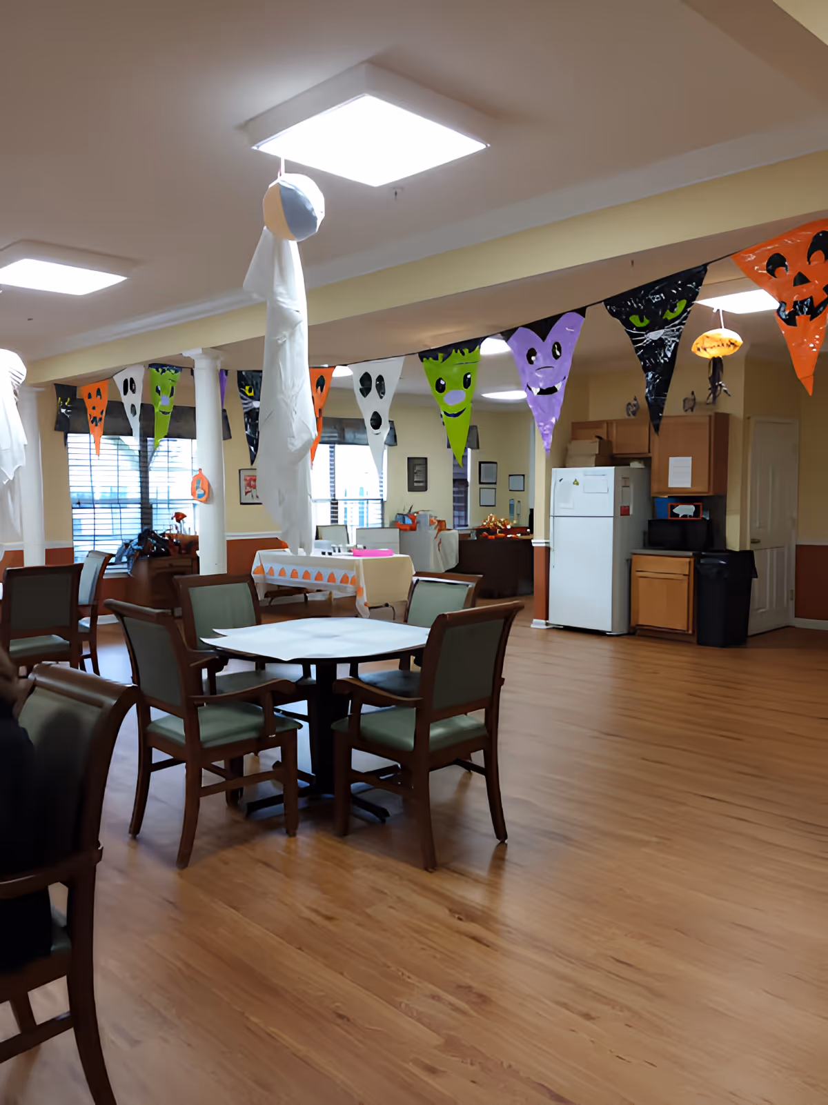 A dining room decorated for Halloween with colorful banners, ghost decorations, and a table set for festivities. The room features wooden flooring and a cozy atmosphere.
